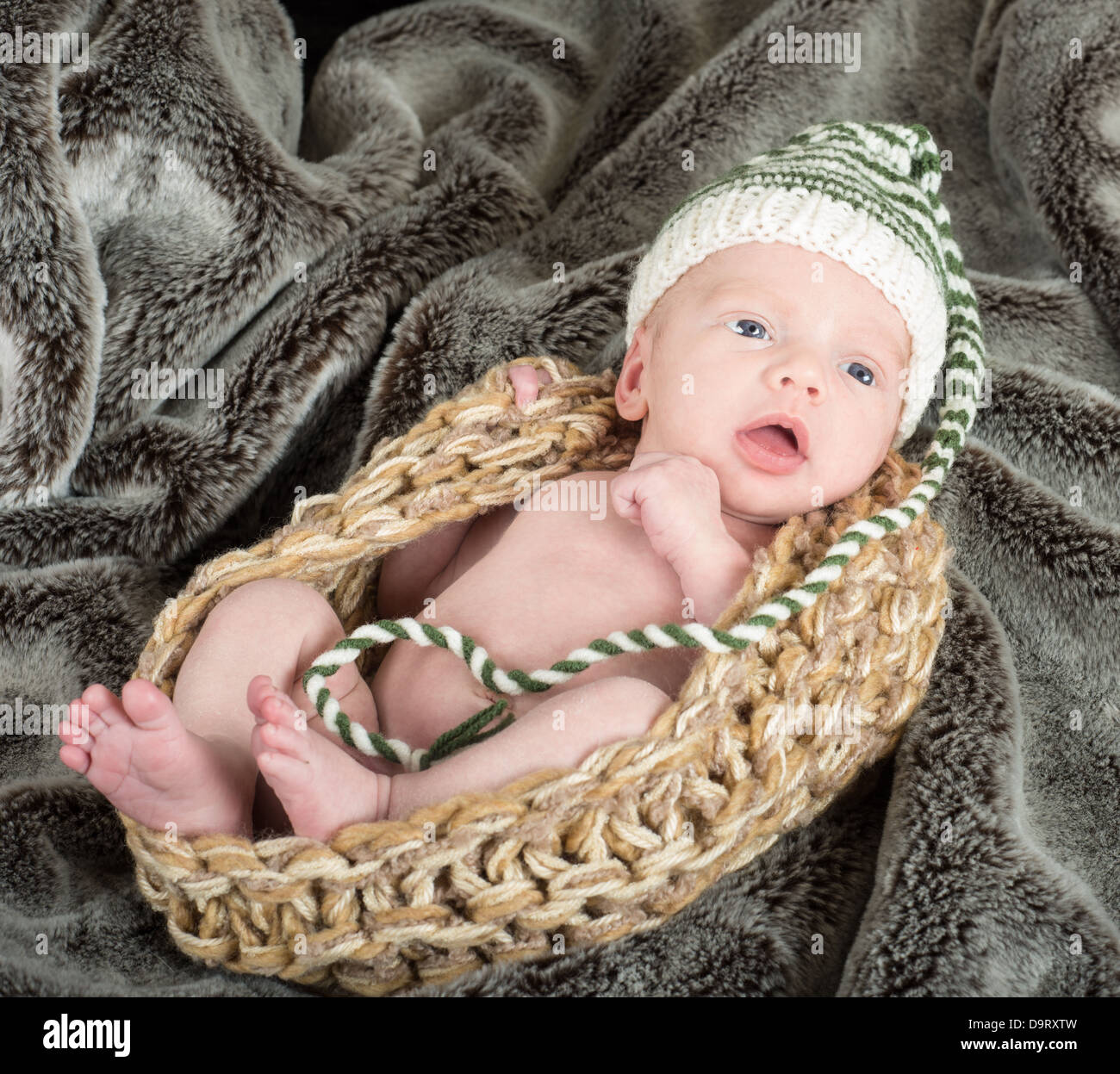 cute baby in a knit basket wearing a green and white knit hat on a fur blanket Stock Photo