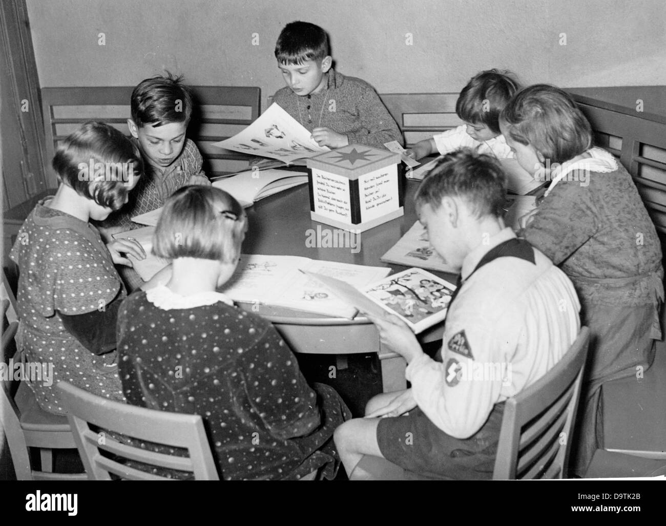 Girls and boys sit at a table and read books in the children's reading room of a library in Berlin, Germany. One boy wears the uniform of the German Youth with a simple Sieg rune on his sleeve. Date unknown. The Nazi Propaganda! on the back of the image reads: 'To the coming week of the German book. A children's reading room and its small regulars. From 25 October to 1 November, the week of the German book will take place this year, whose goal it is to bring together book and people. - Archive image: View of a children's reading room in Berlin: The youngest generation sits peacefully at the ro Stock Photo