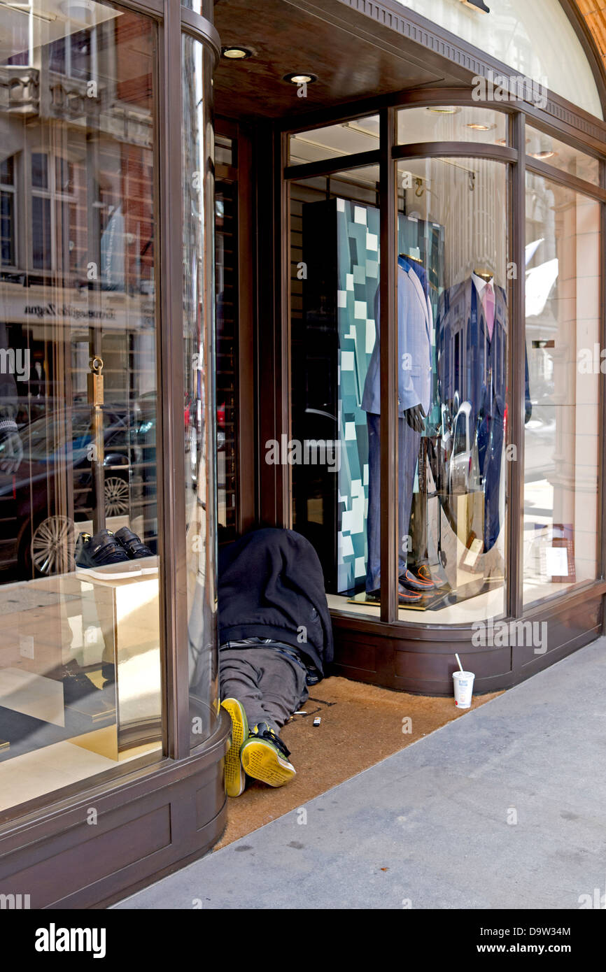 A homeless person is slumped in a doorway of an upmarket clothes store. Portrait view. Stock Photo