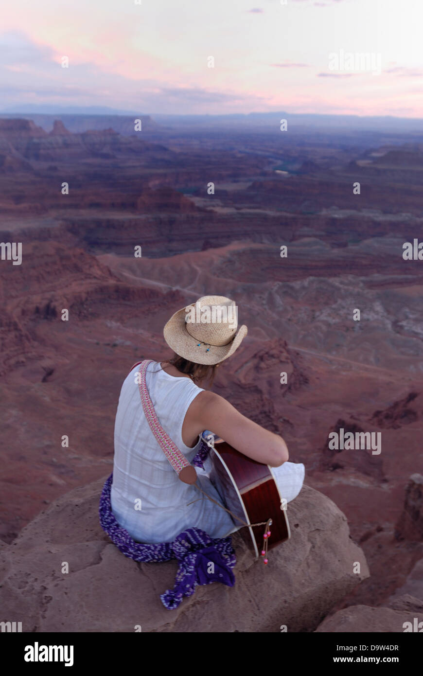 Woman cowboy hat playing guitar and singing song while sitting on edge canyon at Dead Horse Point State Park near Moab Utah USA Stock Photo