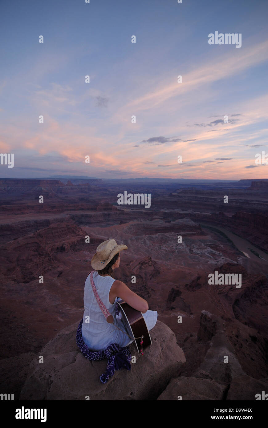 Woman cowboy hat playing guitar and singing song while sitting on edge canyon at Dead Horse Point State Park near Moab Utah USA Stock Photo