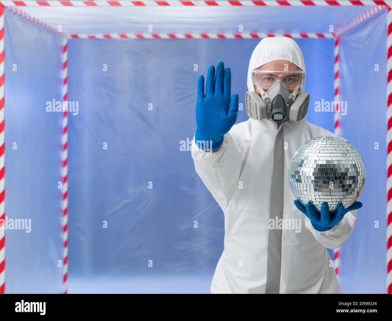 person dressed in a bio hazard protective suit making a halt gesture with one hand and holding a disco ball in the other in front of a confinement tent Stock Photo