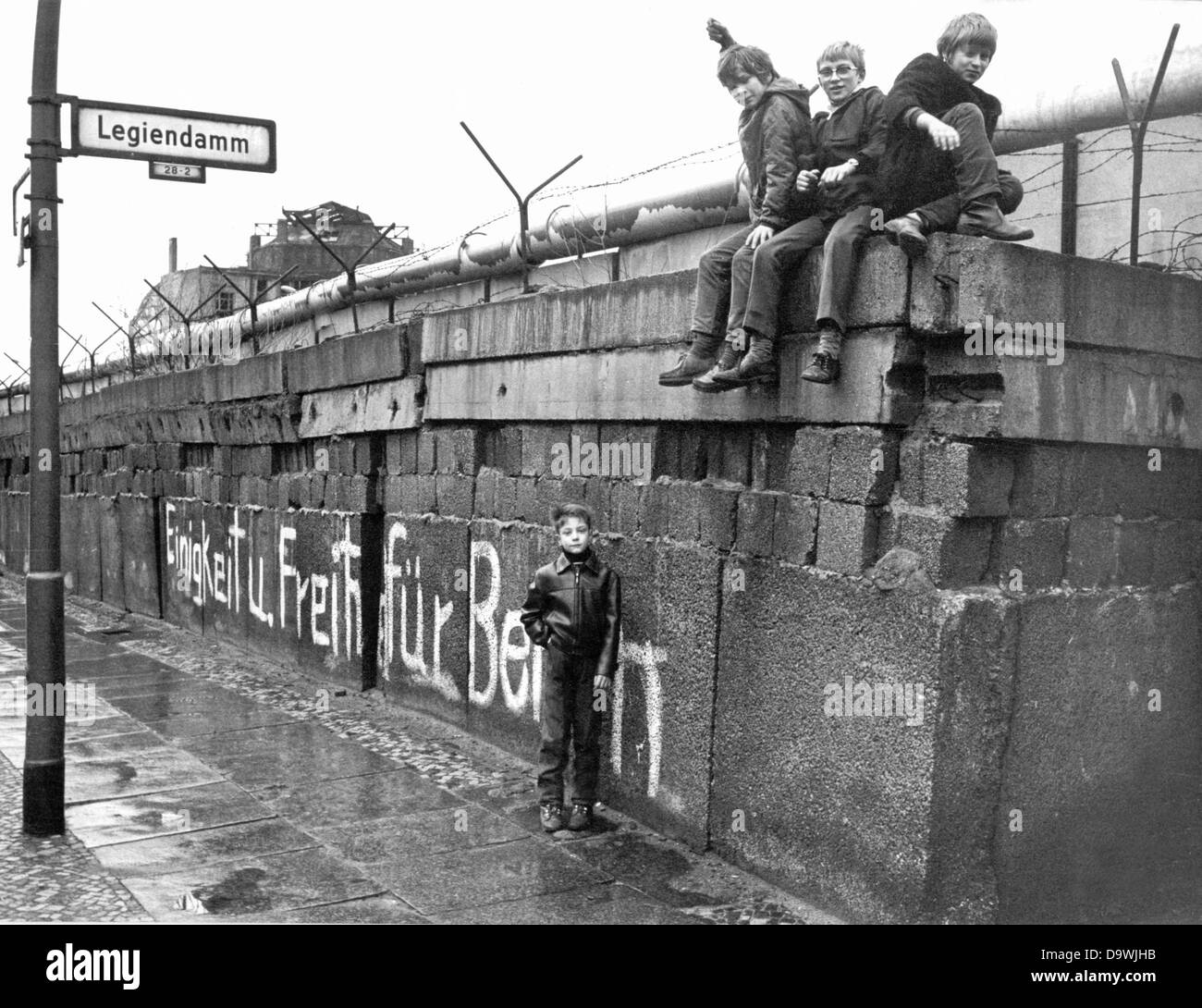 A group of children sits on the Berlin Wall at the 'Legiendamm' in the West Berlin borough Kreuzberg, March 1972. A white graffiti translates 'Unity and Freedom for Berlin'. Stock Photo