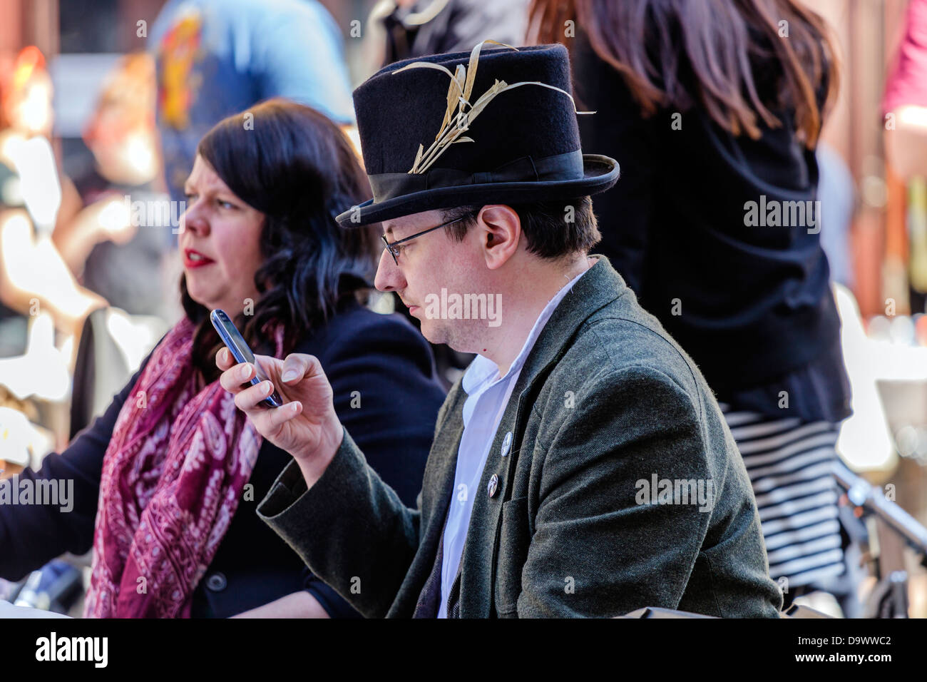 Man with a top hat sitting at a table outside a pub, checking his phone Gibson Street, West End Festival, Glasgow, Scotland, UK Stock Photo