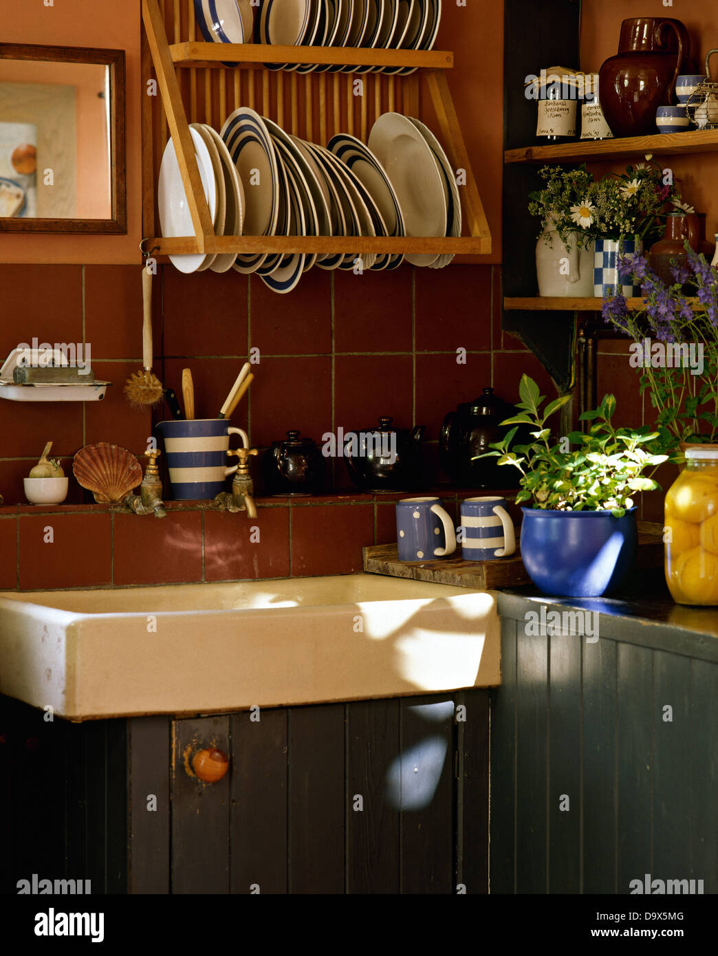 Blue+white striped plates on wooden rack above Belfast sink in cottage kitchen with brown tiles and blue painted units Stock Photo