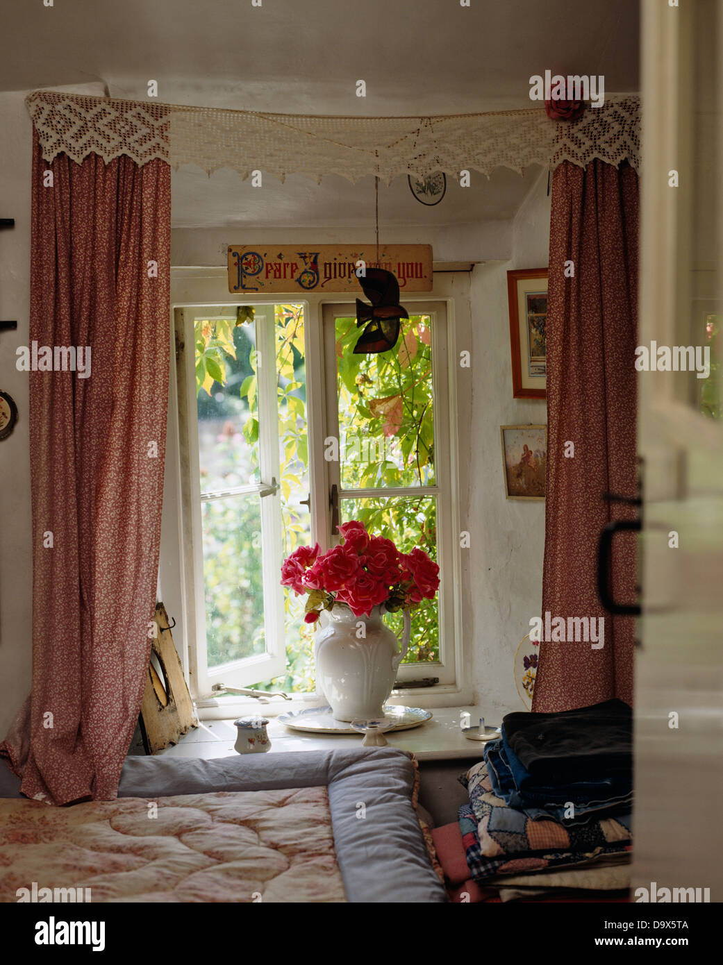 Pink curtains with lace pelmet on open window with vase of red roses on windowsill in cottage bedroom Stock Photo