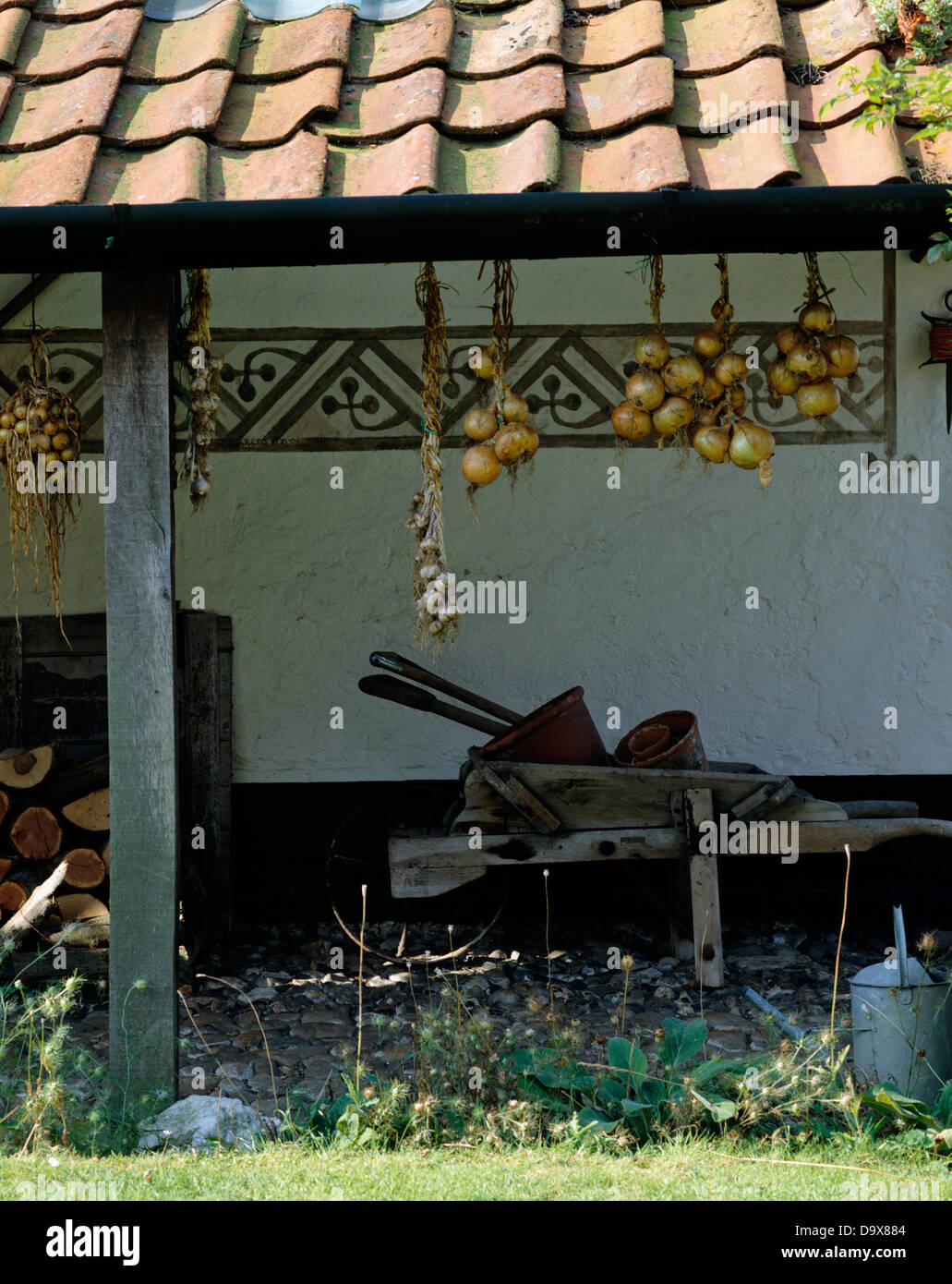 Old wooden wheelbarrow on covered veranda with onions hanging from red tiled roof Stock Photo