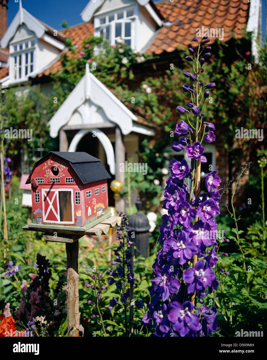 Close-up of red painted bird house beside blue delphinium in cottage garden in summer Stock Photo
