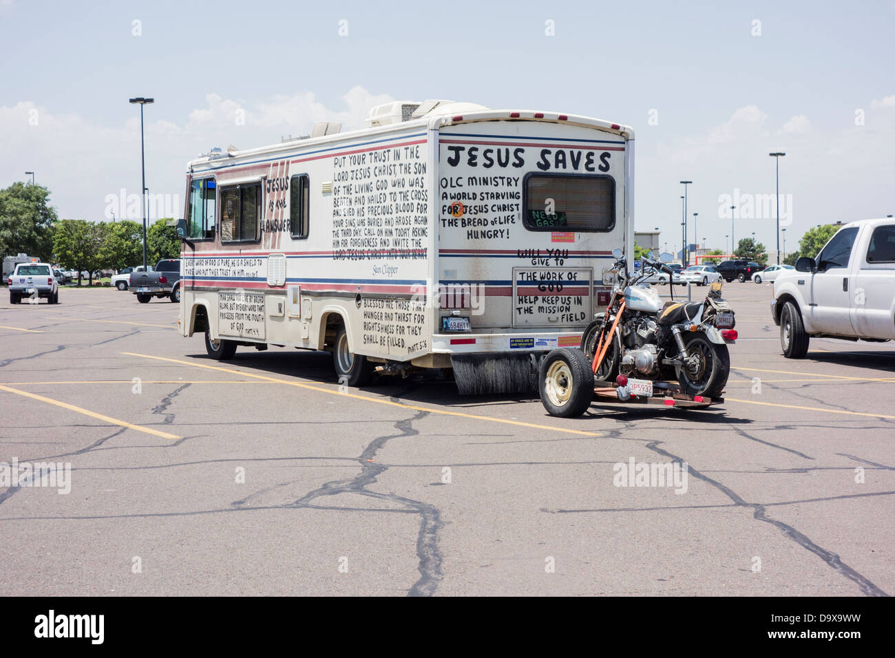 An old recreational vehicle towing a motorcycle and hand-painted with biblical scripture sits in a parking lot. Stock Photo