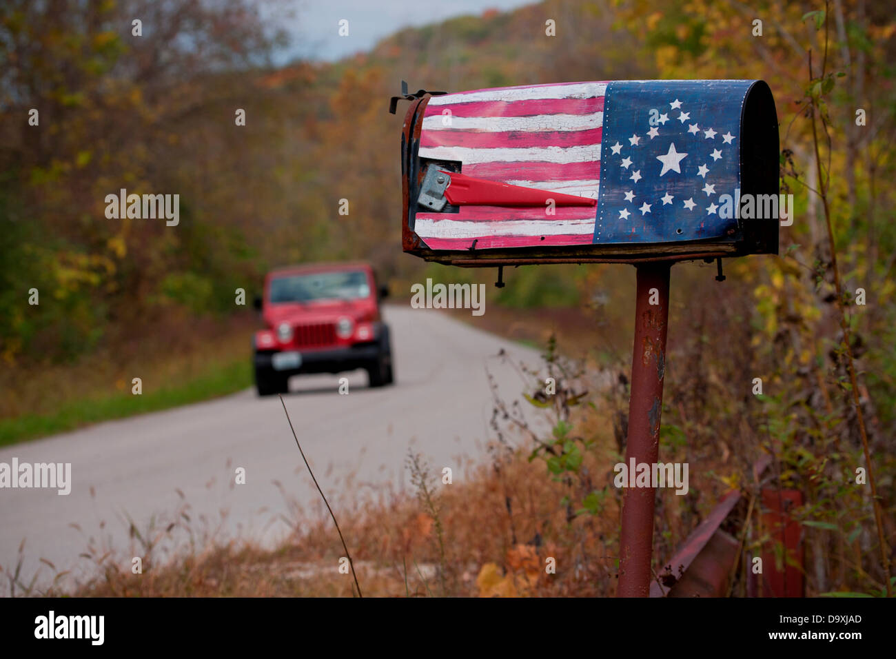 Red Jeep in background drives by weathered, mailbox with US Flag painted on the side of it Stock Photo