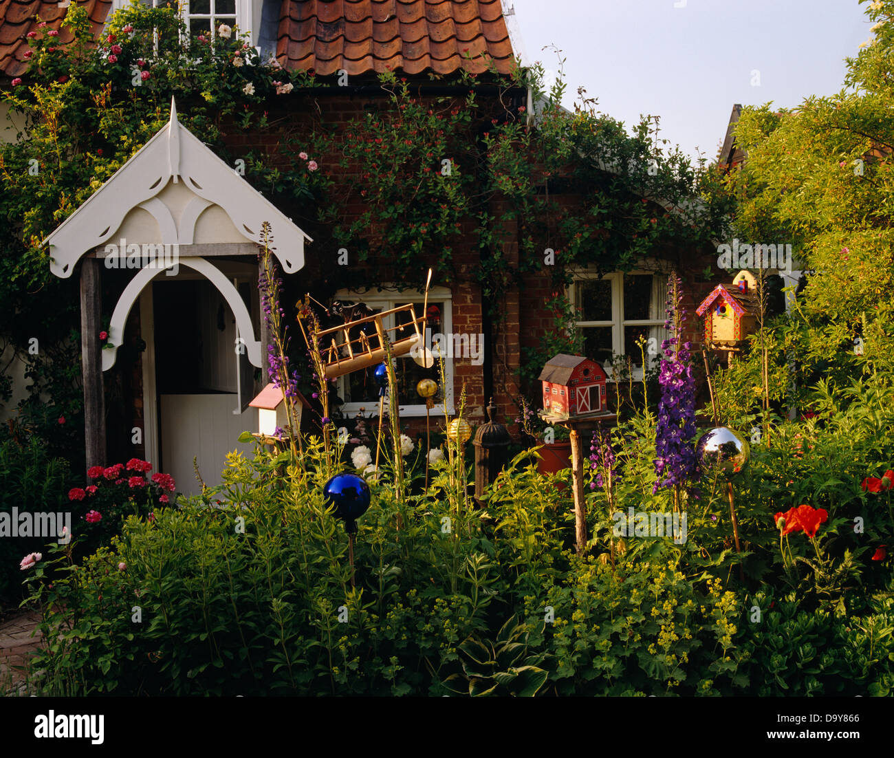 Wooden bird houses on sticks among flowering summer plants in garden of country cottage with white porch Stock Photo