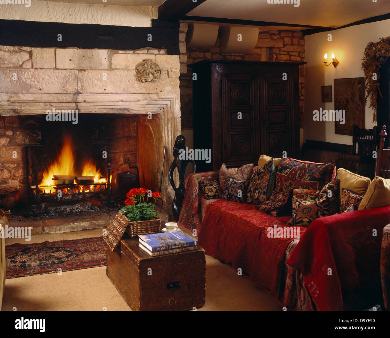 Red throw on sofa piled with cushions beside fireplace with open fire