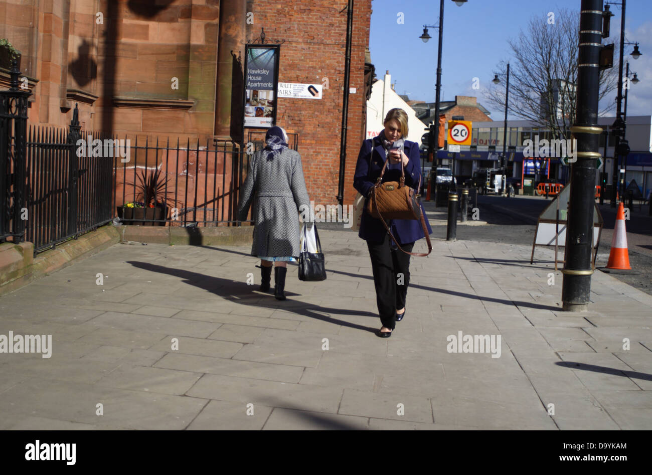 Young woman walking towards cam looking in handbag. Old lady walking away from cam, carrying handbag/shopping bag. Street  scene Stock Photo