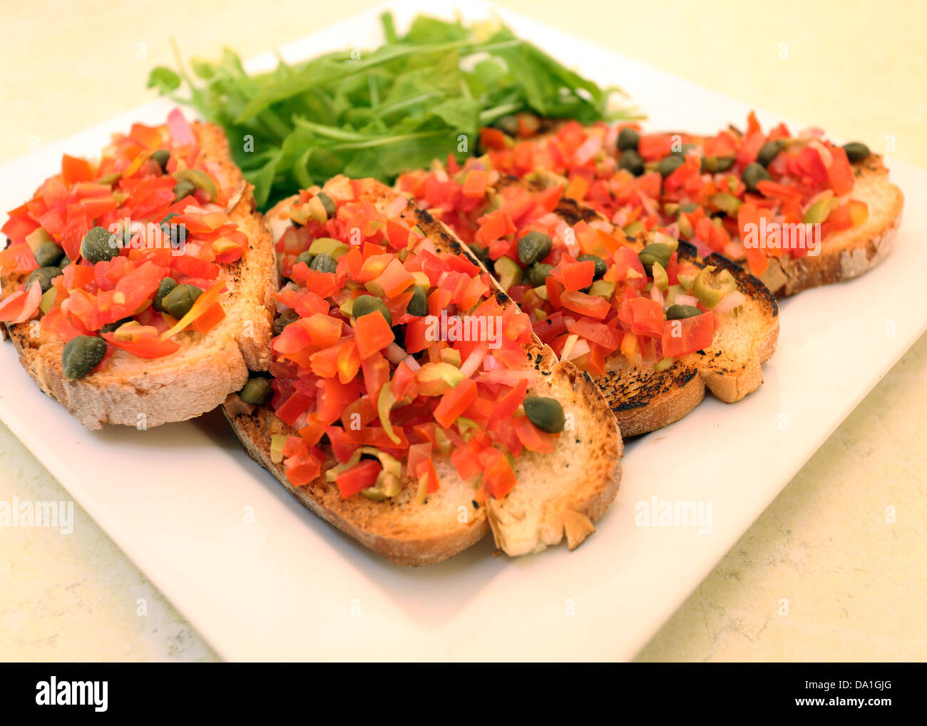 SQUARE PLATE WITH BRUSCHETTA: SLICES OF CRISPY BREAD TOPPED WITH CHOPPED TOMATOES,GARLIC,OLICE OIL AND CAPERS,ROCKET LEAVES GARN Stock Photo