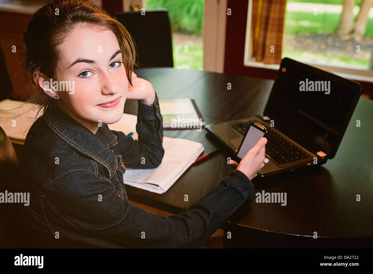 Teenaged girl is studying at the family kitchen table and researches something on her phone Stock Photo