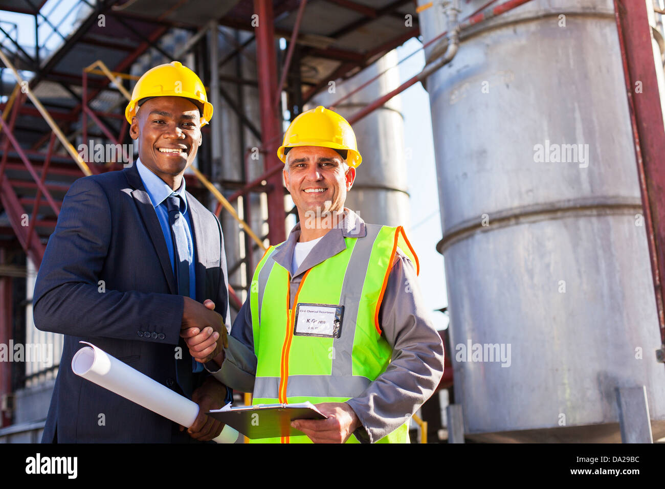 cheerful african refinery manager handshaking with senior worker in front of fuel-tanks Stock Photo