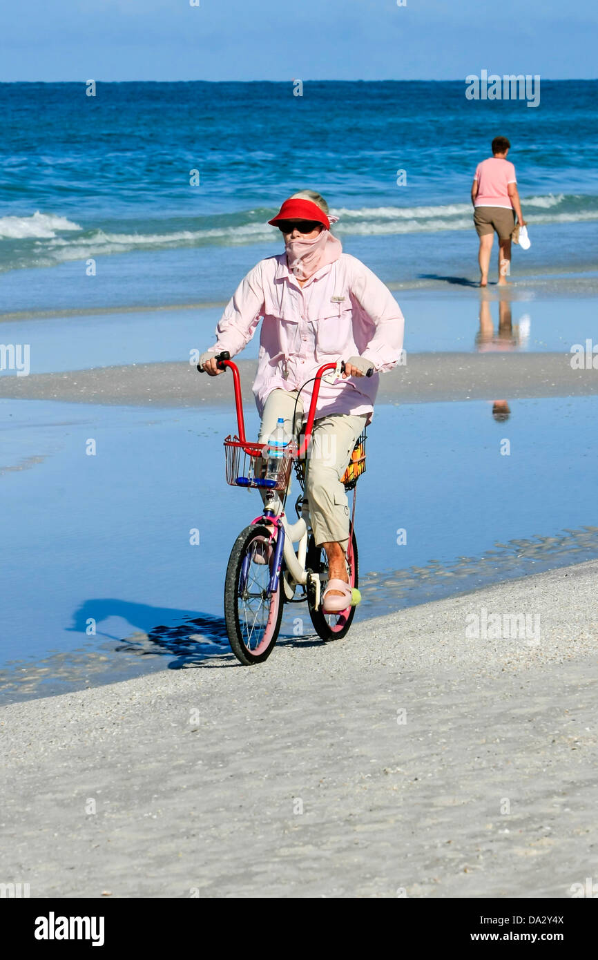 Adult woman wearing clothes to guard against the sun as she rides her bicycle on the beach in Florida Stock Photo