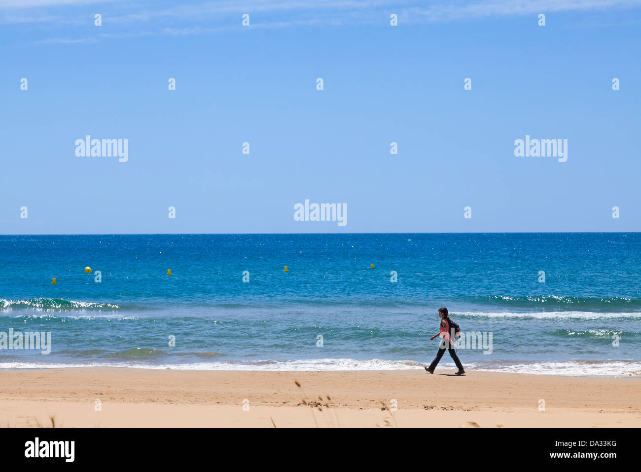 one woman with backpack walking purposefully alond beach by sea Stock Photo