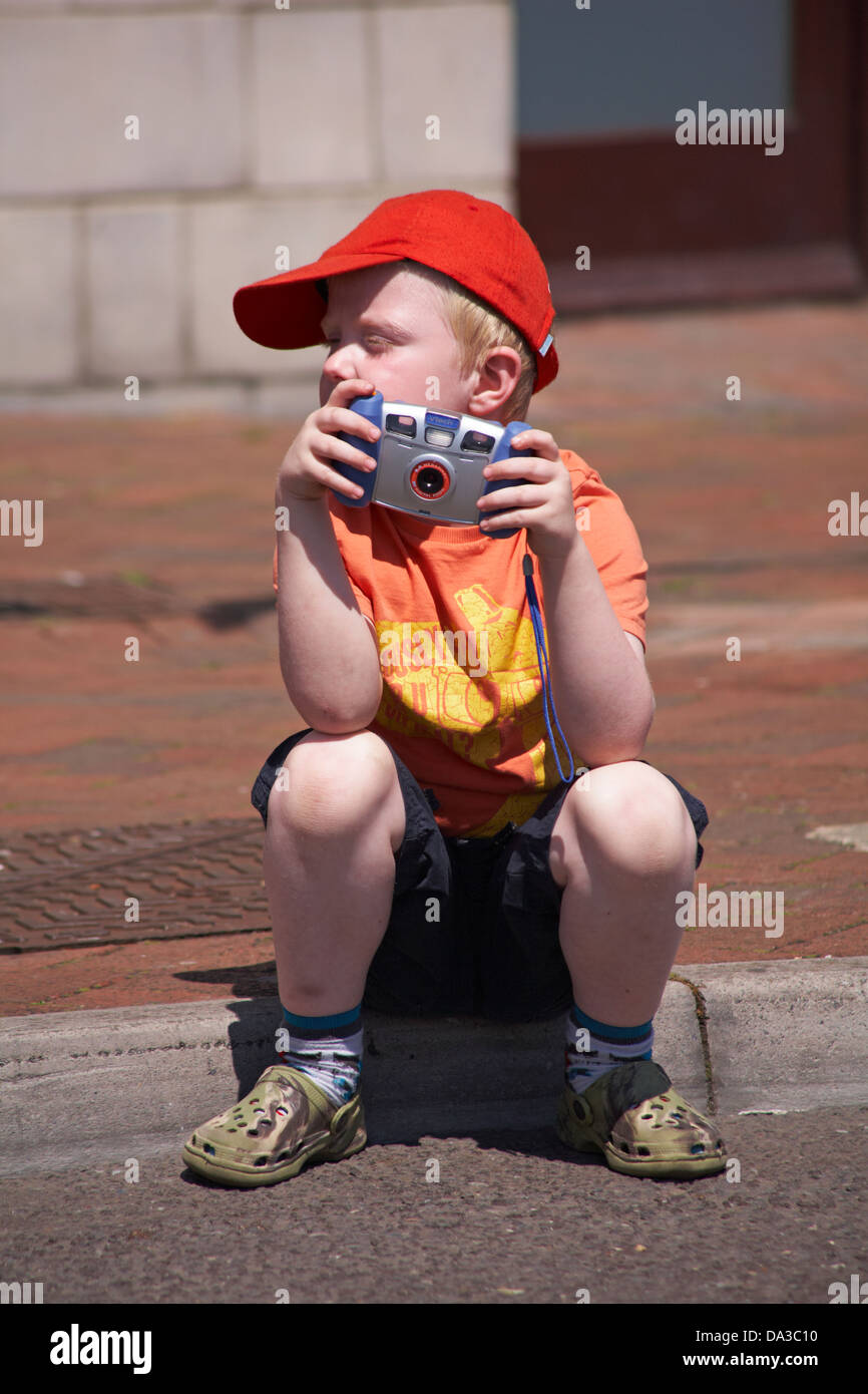 young boy sitting on pavement holding vtech digital zoom camera watching festival at Poole in June Stock Photo