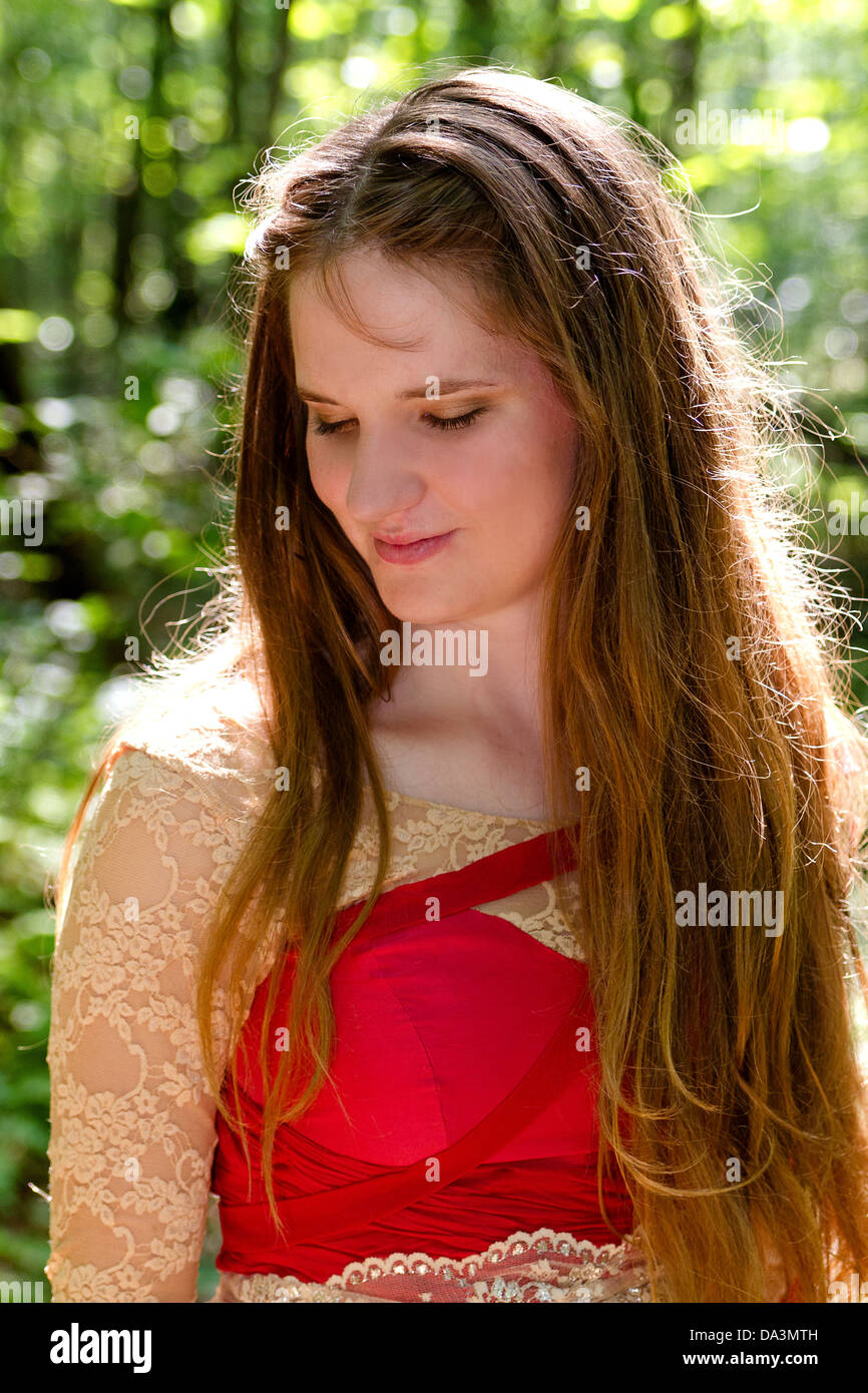 Beautiful young woman with long golden-brown hair smiles at ground in woods, dressed in red medieval gown with lace sleeves Stock Photo