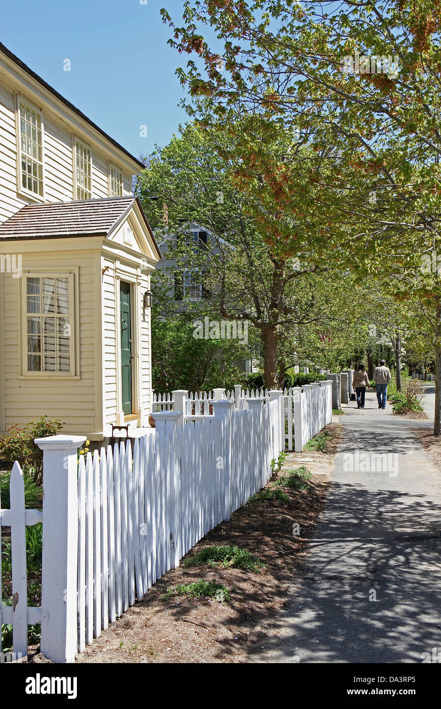 A yellow home behind a white picket fence, in Concord, Massachusetts, two people walking on the sidewalk in the distance. Stock Photo