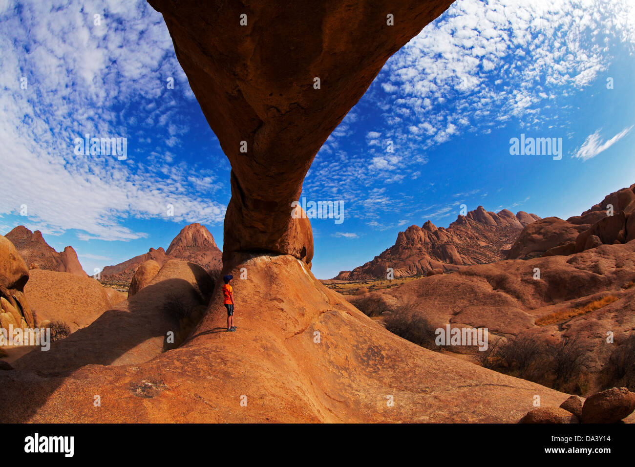 Boy under natural rock arch at Spitzkoppe (left), and Pondok Mountains in distance (right), Namibia, Africa Stock Photo