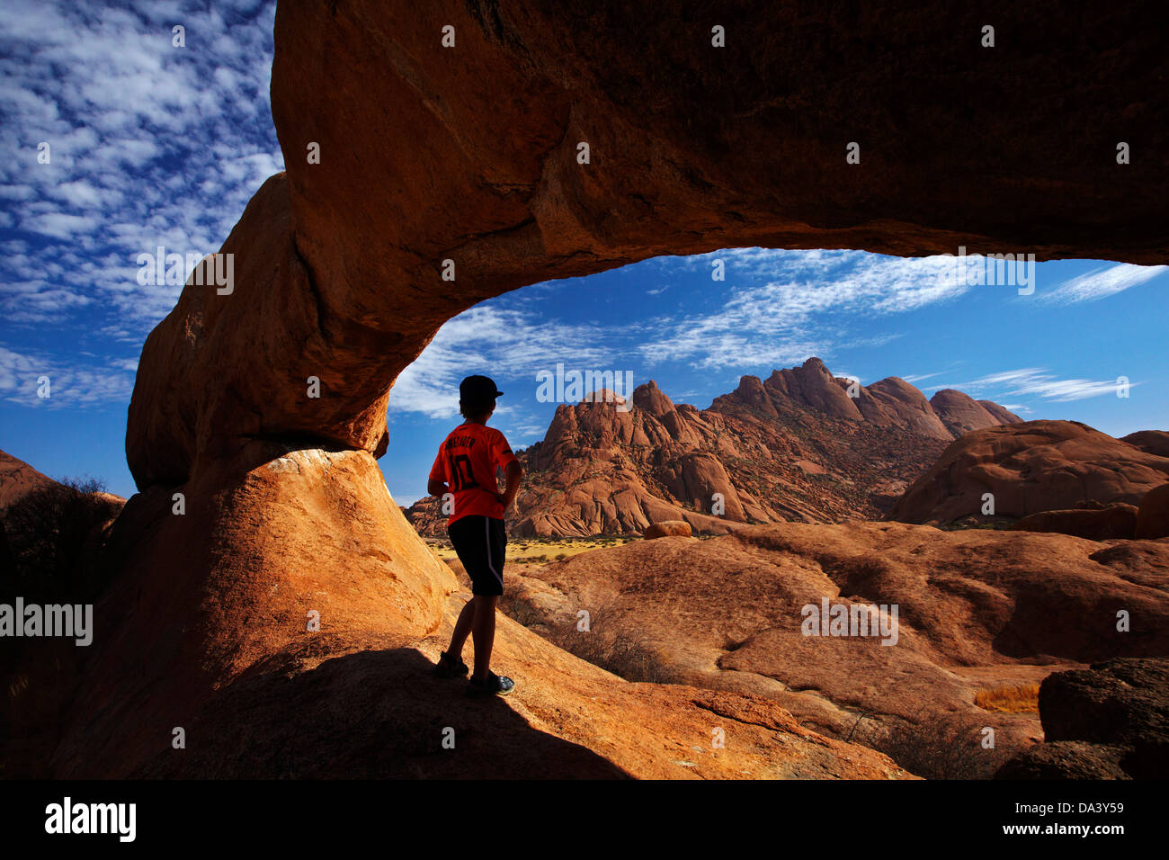 Boy under natural rock arch at Spitzkoppe, and Pondok Mountains in distance, Namibia, Africa Stock Photo