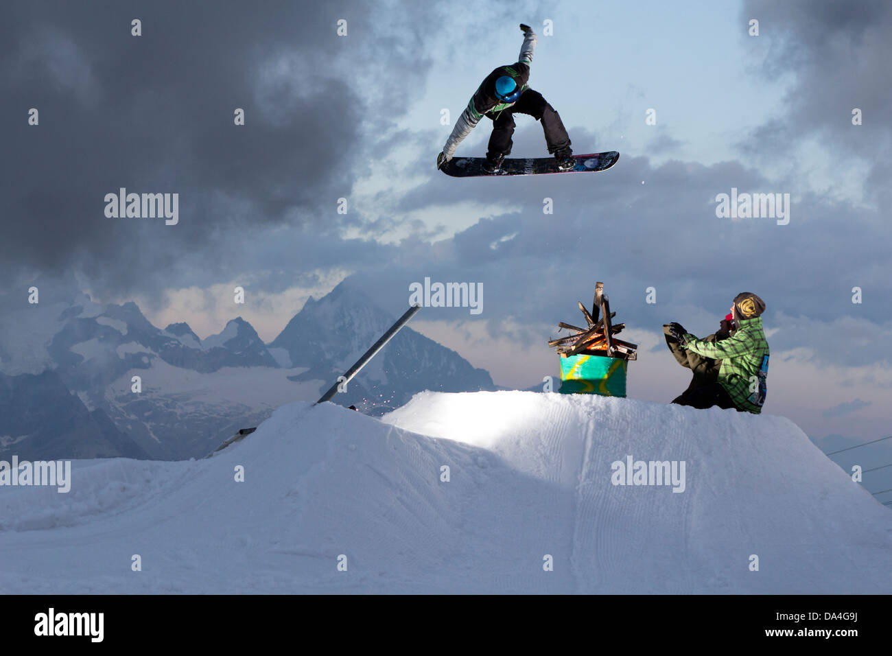 ZERMATT, SWITZERLAND. A freestyle snowboarder is executing a tailgrab trick and jumps over an oil barrel with a fire on it. Stock Photo