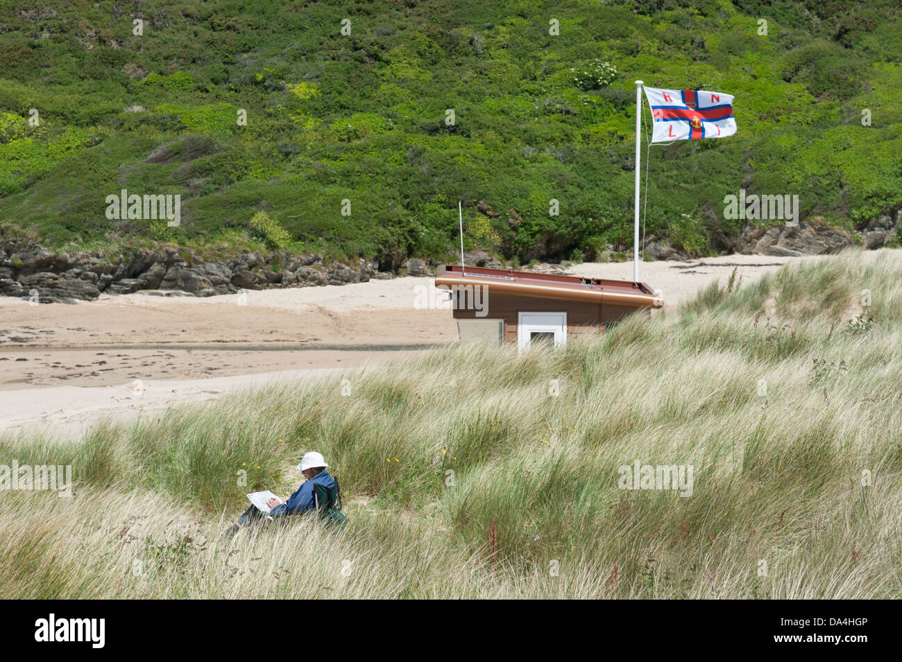 Man sitting in a deckchair in the dunes reading a paper by the RNLI lifeguard hut at Porthcothan Bay Cornwall UK Stock Photo