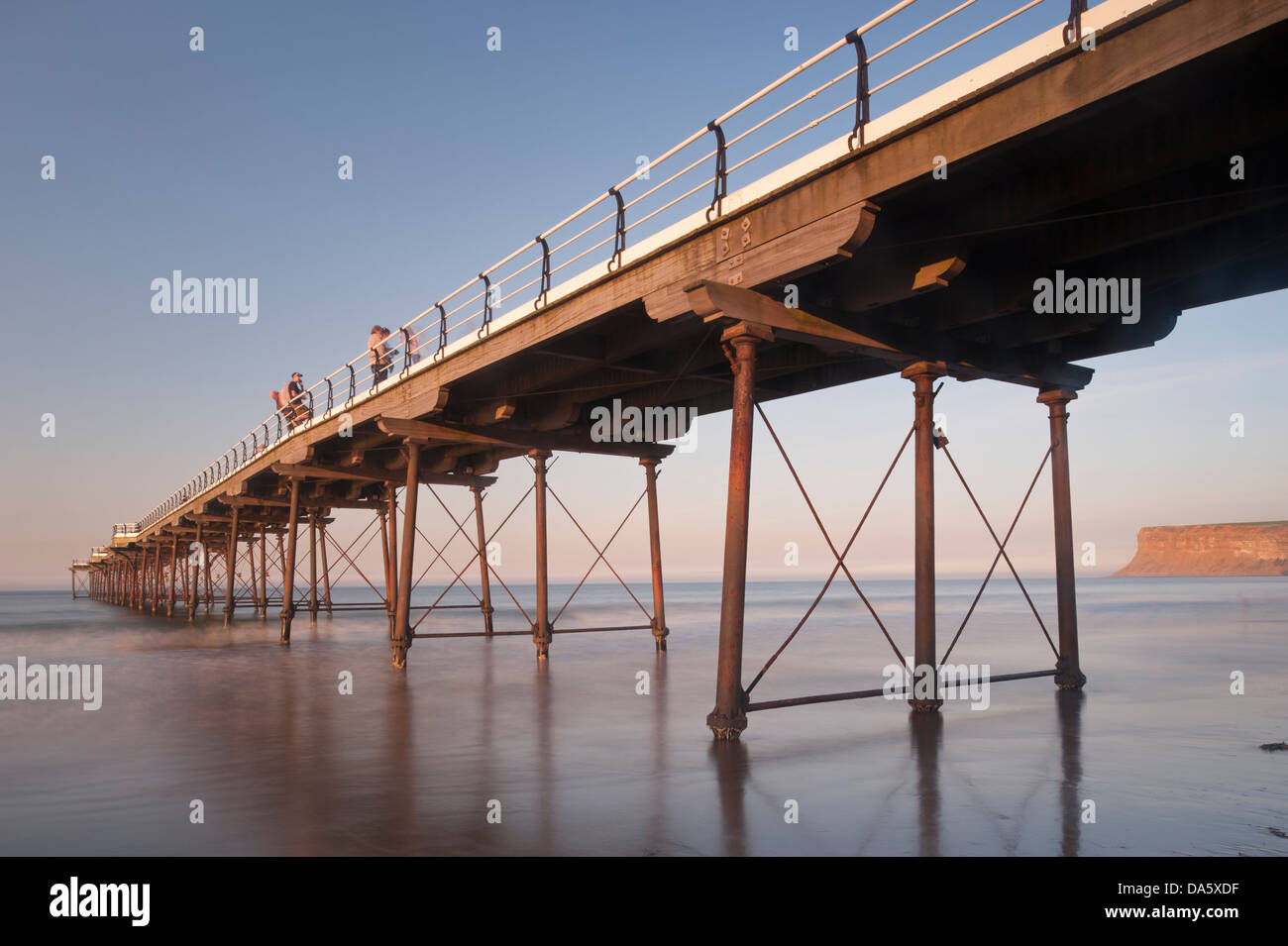 Blue summer evening sky & view of people standing & walking in sun, on historic scenic seaside pier over calm sea - Saltburn-by-the-Sea, England, UK. Stock Photo