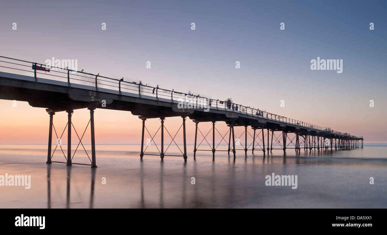 Under colourful summer sunset sky, view from sandy beach of people walking on historic seaside pier over calm sea - Saltburn-by-the-Sea, England, UK. Stock Photo