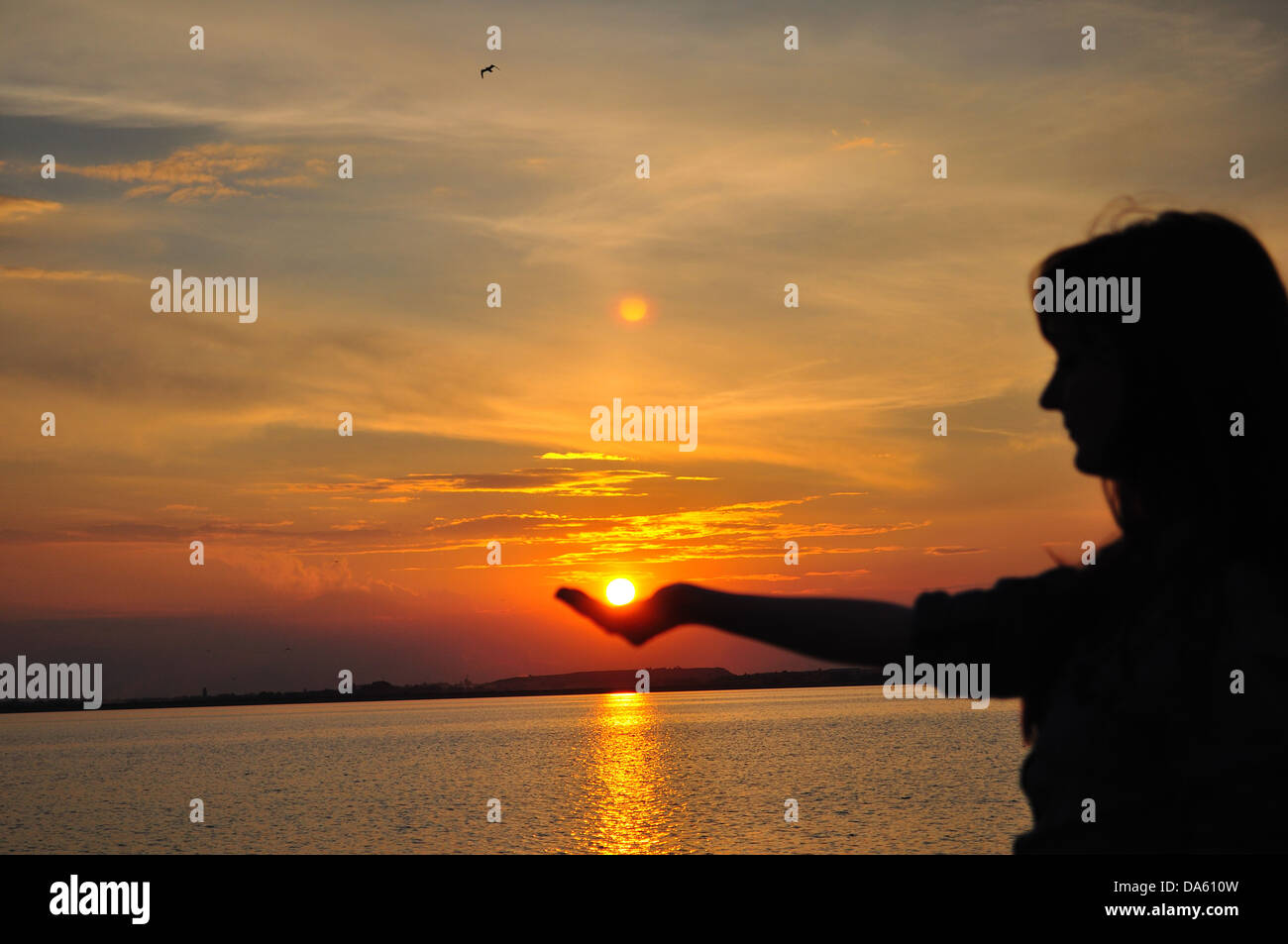 Woman holding the sun in hand at sunset next to a lake Stock Photo - Alamy