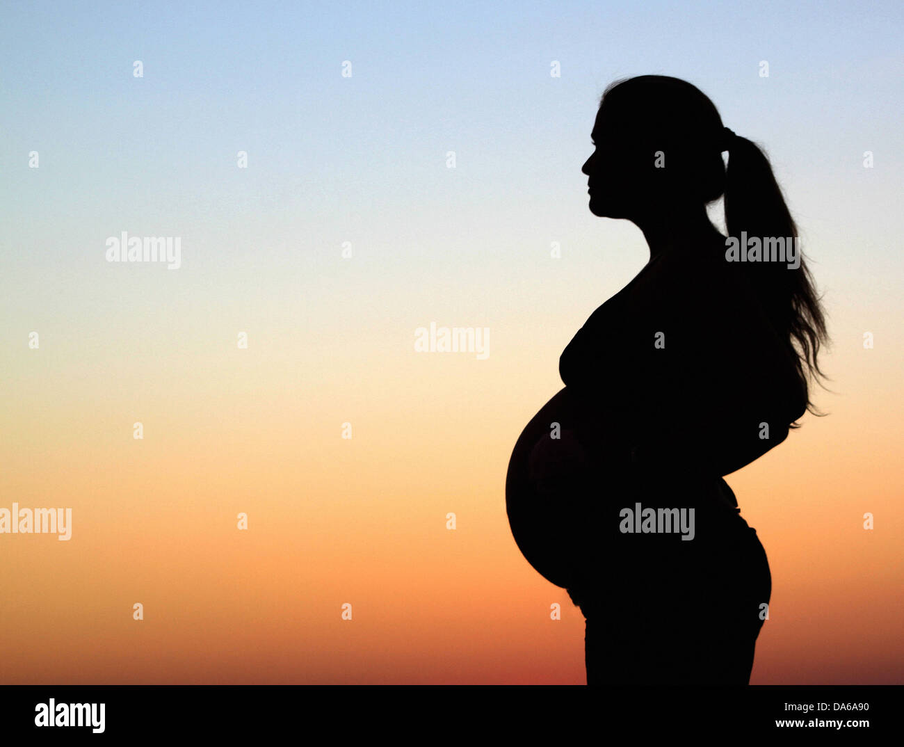 A side profile silhouette of a pregnant woman standing in front of a warm orange and blue sky Stock Photo