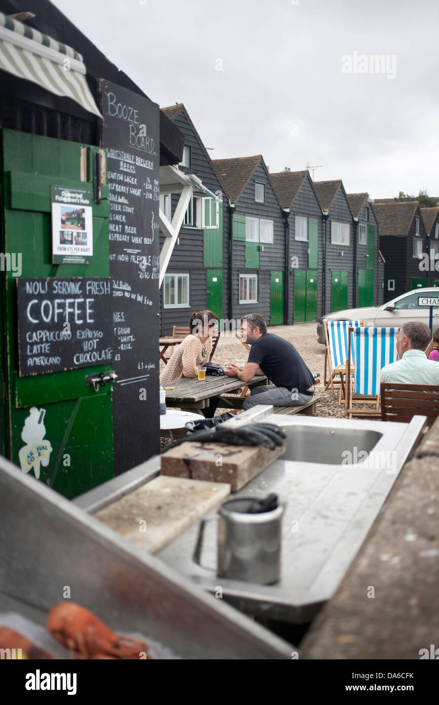 People seated at picnic table outside cafe hut near Whitstable beach with converted fishing huts with green doors in background Stock Photo