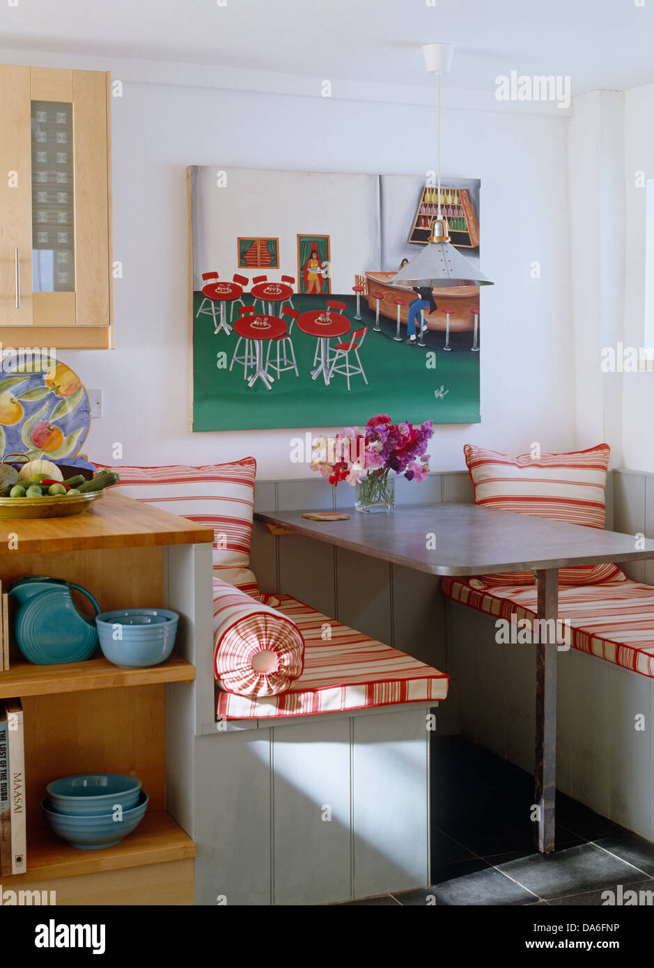 Colorful painting on wall behind fixed metal table and built in seating with red striped cushions in corner of country kitchen Stock Photo