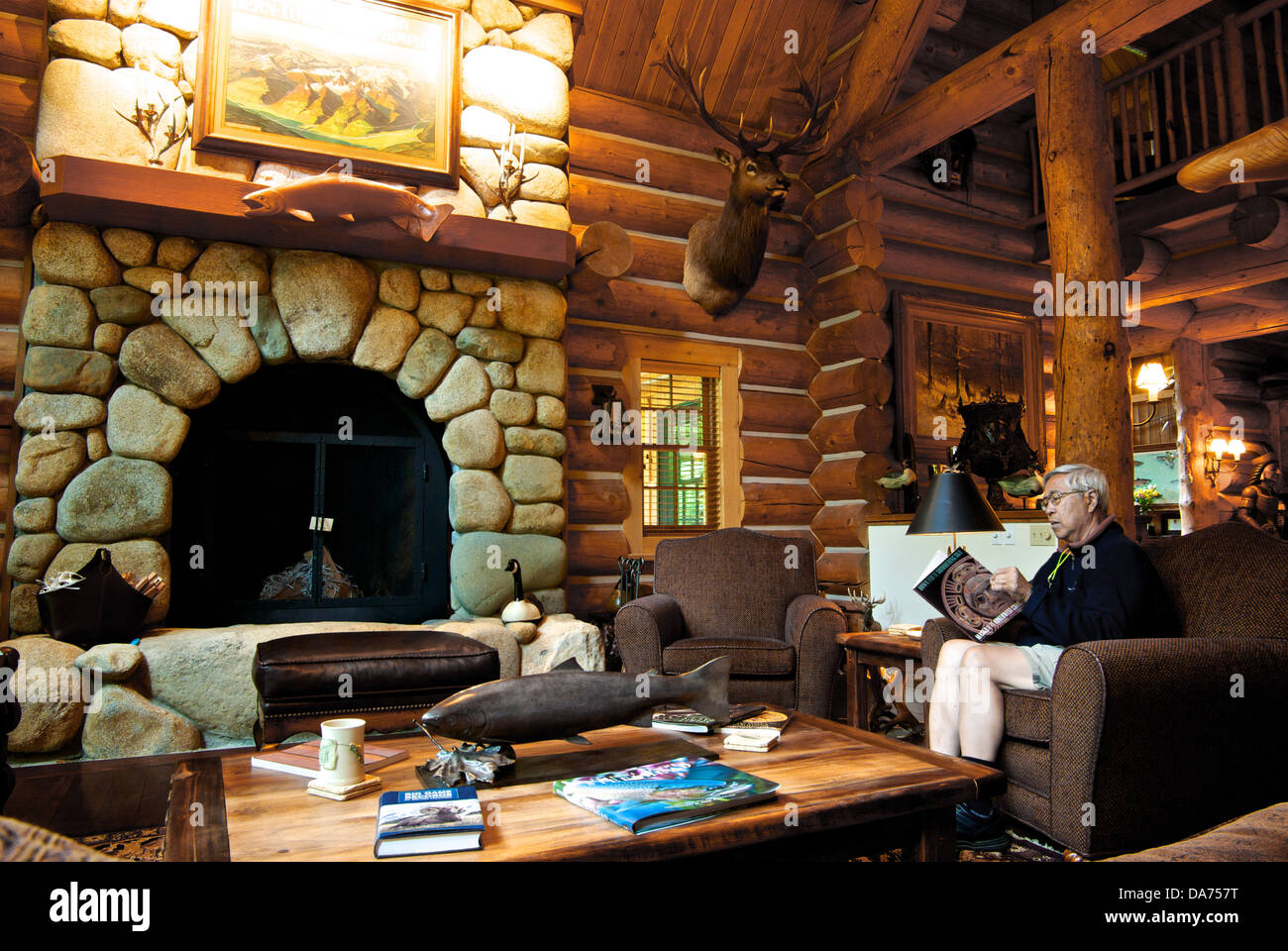 Asian man reading log construction wood plank walls stone fireplace great room Gold River Lodge BC Stock Photo