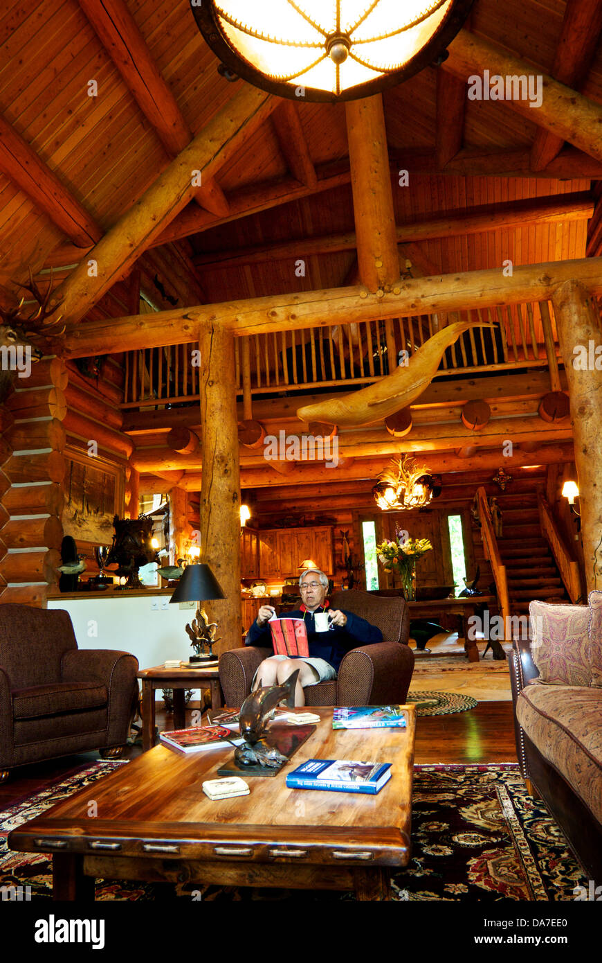 Asian man reading log beam construction wood plank walls stone fireplace great room Gold River Lodge BC Stock Photo