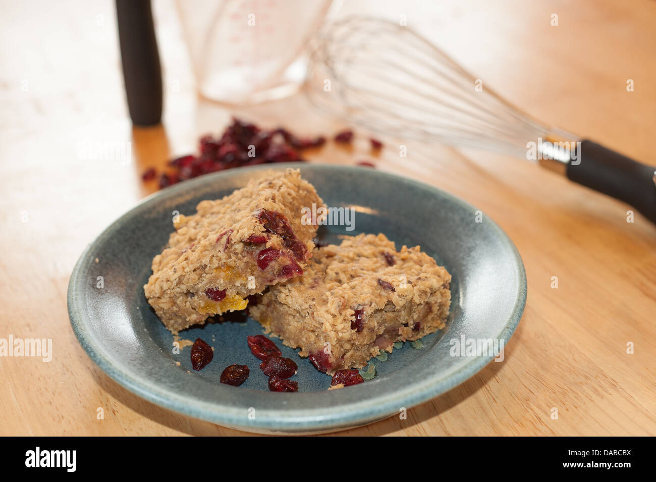 Color images of homemade oat and cranberry energy bars on a pottery plate showing some of the ingredients and utensils on table. Stock Photo