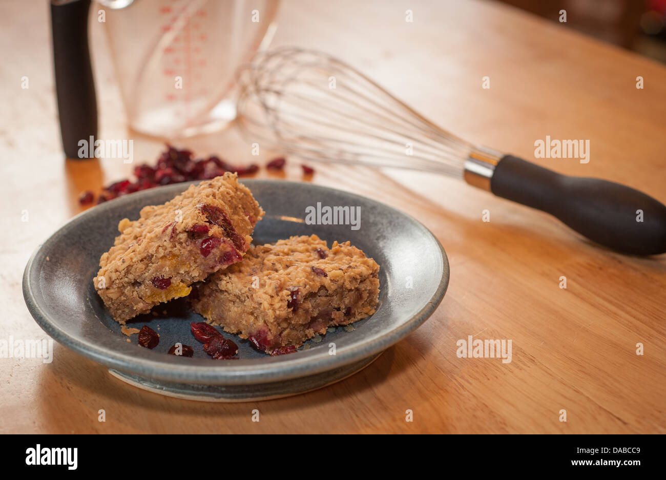 Color images of homemade oat and cranberry energy bars on a pottery plate showing some of the ingredients and utensils on table. Stock Photo