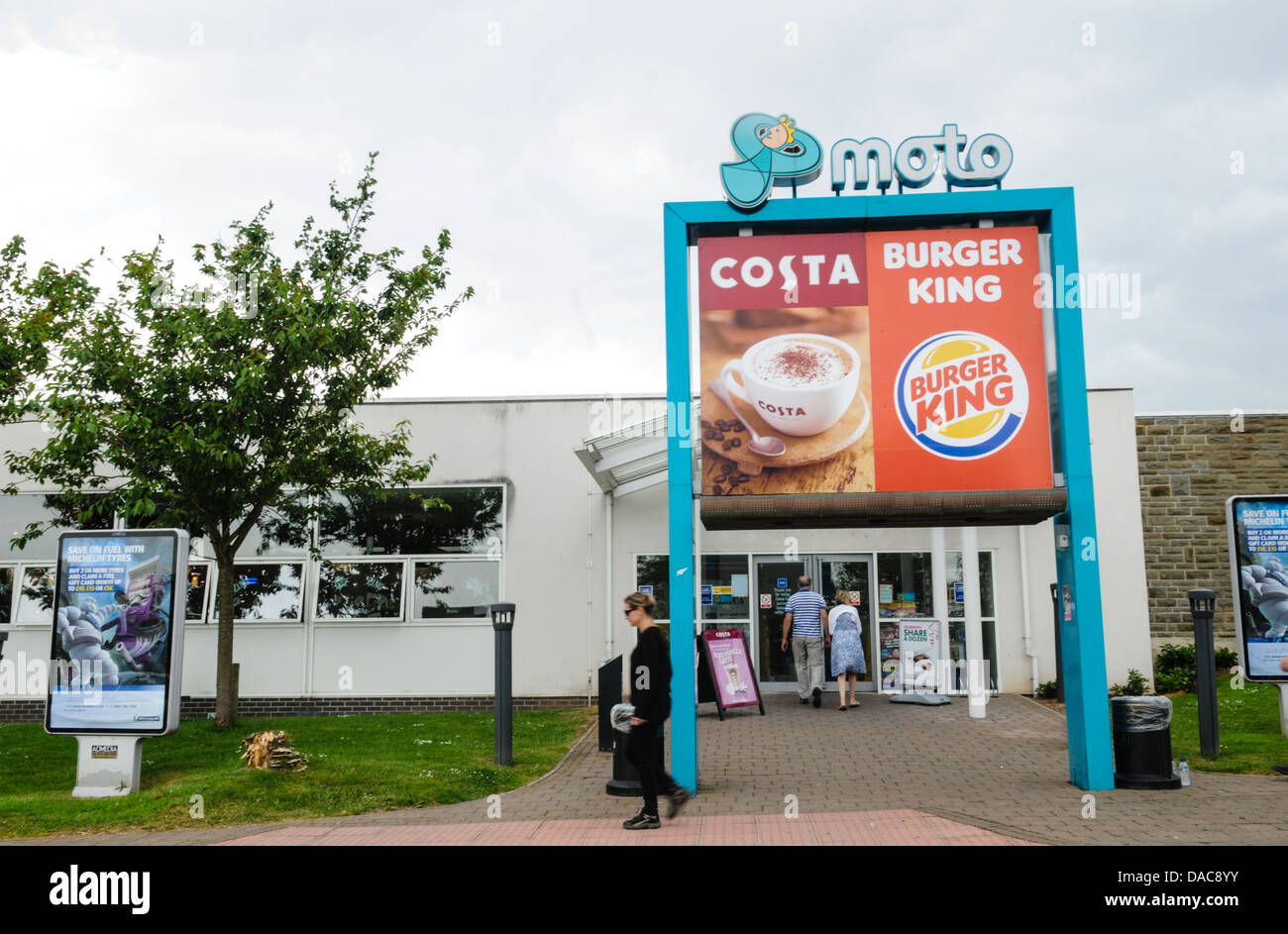 Large Moto, Costa and Burger King sign at the entrance to a motorway service station with a woman walking past at Aust Services Stock Photo