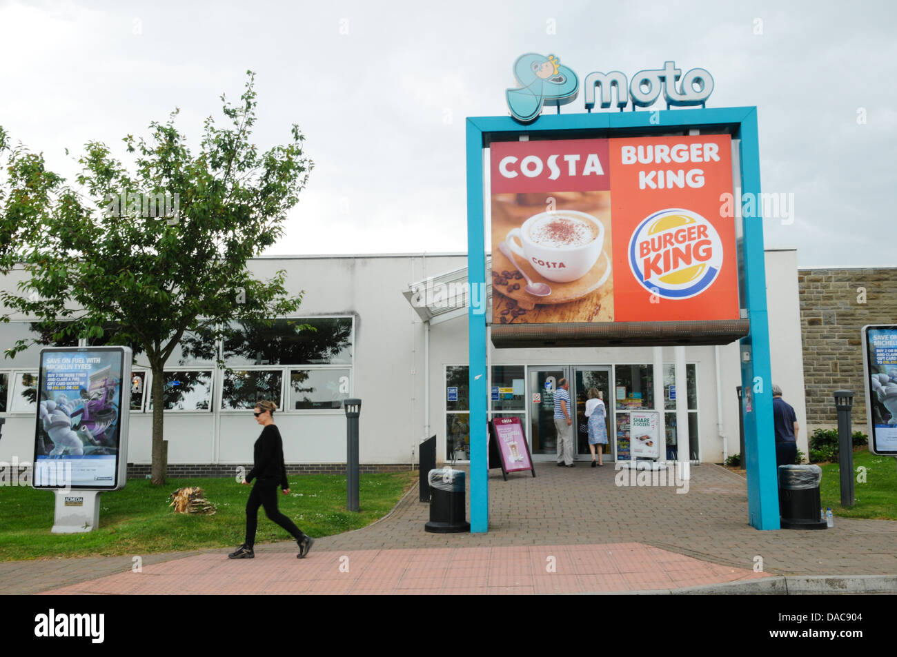 Large Moto, Costa and Burger King sign at the entrance to a motorway service station with a woman walking past at Aust Services Stock Photo