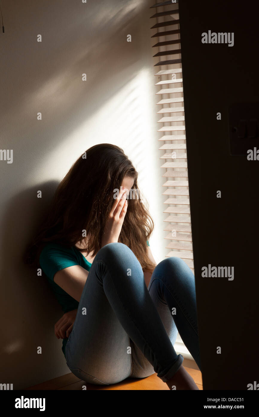 A teenage girl, hand on her face, sitting by a window with light pouring in. Stock Photo