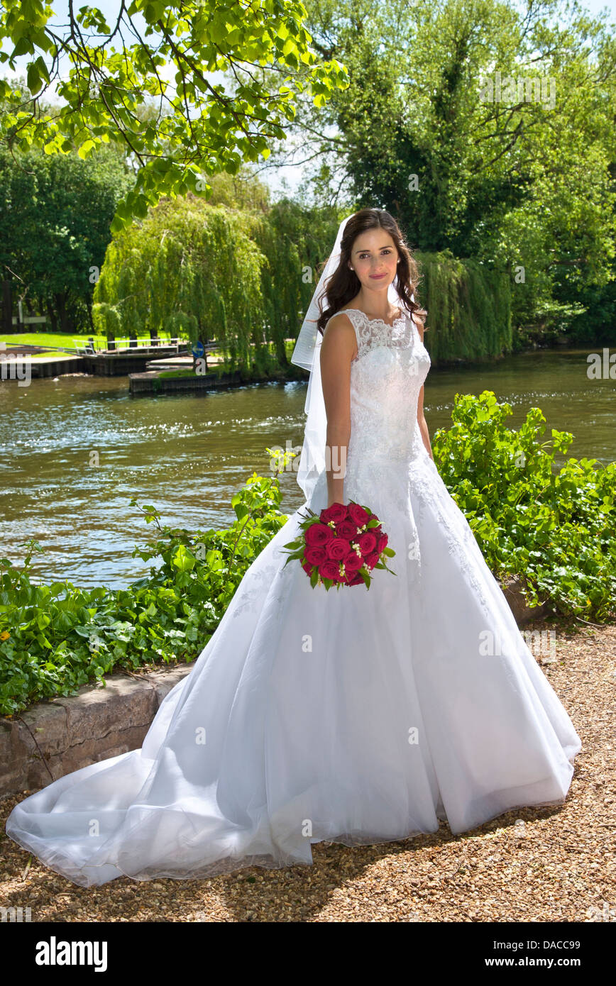 Bride in traditional white bridal dress with red rose bouquet just married, posing in riverside church grounds Stock Photo