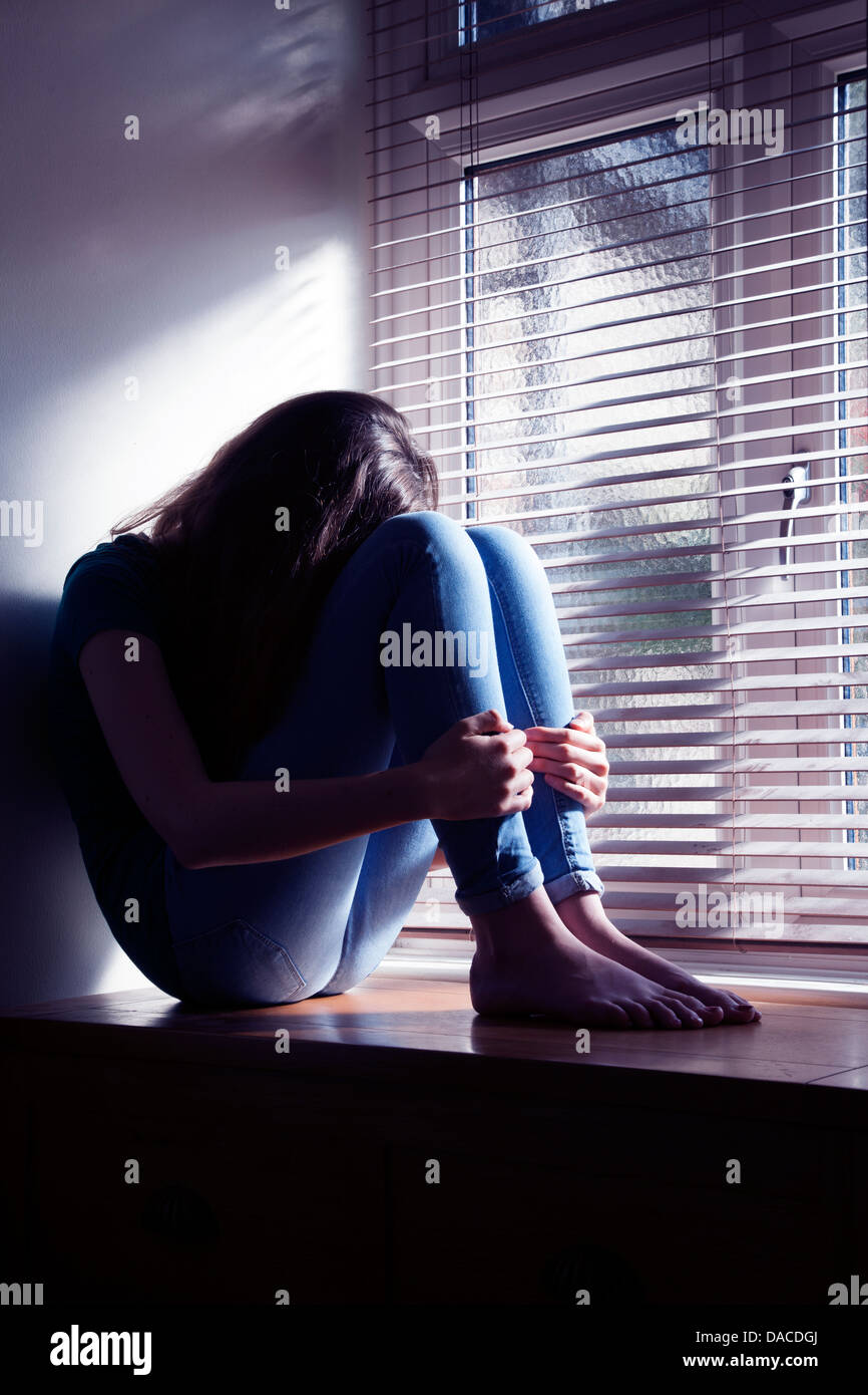 Teenage girl, head bowed, sitting by a window with light pouring in. Stock Photo