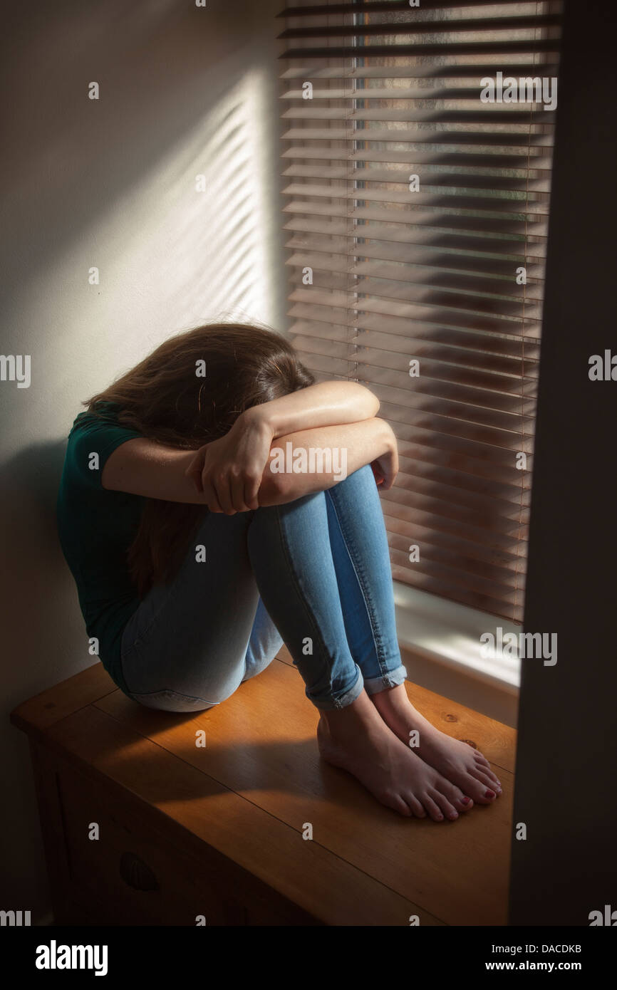 A teenage girl, head down, sitting by a window with light pouring in. Stock Photo