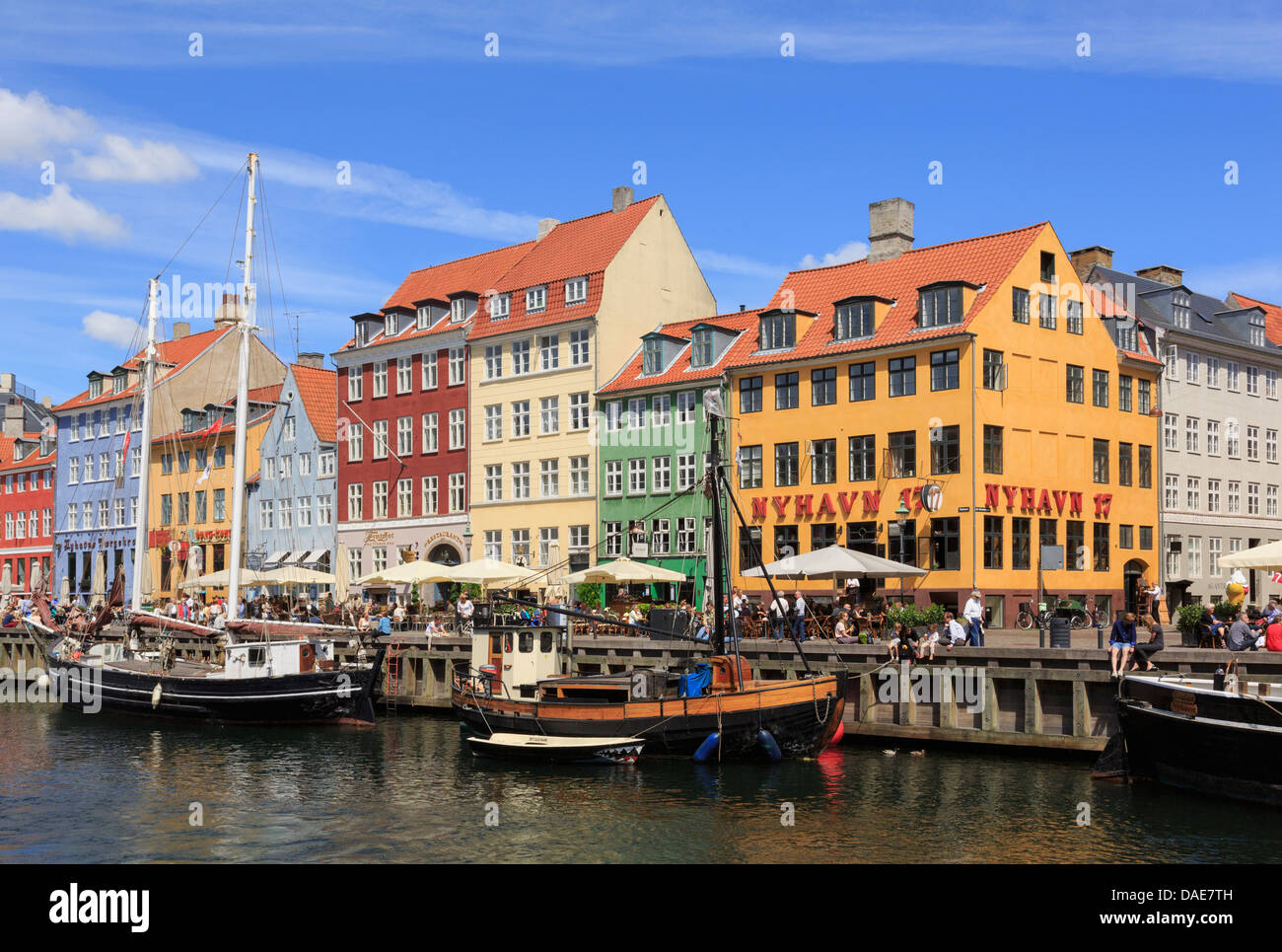 Old wooden boats moored on canal by colourful 17th century buildings on waterfront busy with people in old city. Nyhavn Copenhagen Denmark Stock Photo