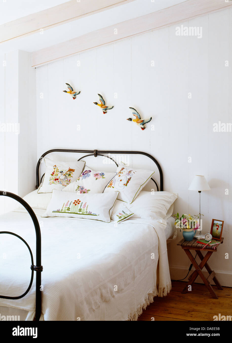 Pottery flying ducks on wall above wrought iron bed with white bed-cover and embroidered cushions in white coastal bedroom Stock Photo