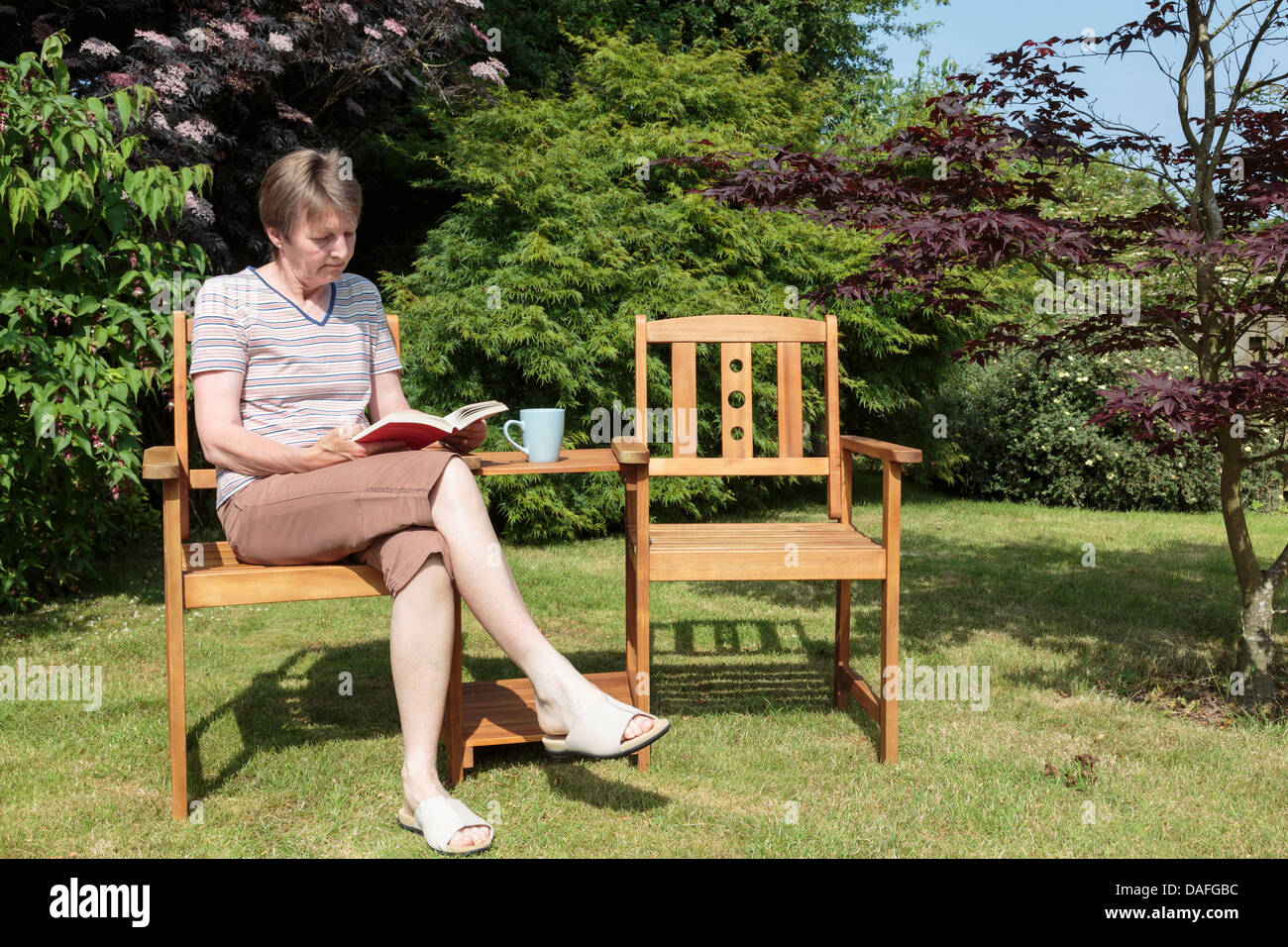 A senior retired woman retiree sitting alone reading a book outdoors relaxing on a seat in a domestic back garden at home on a sunny day in summer UK Stock Photo