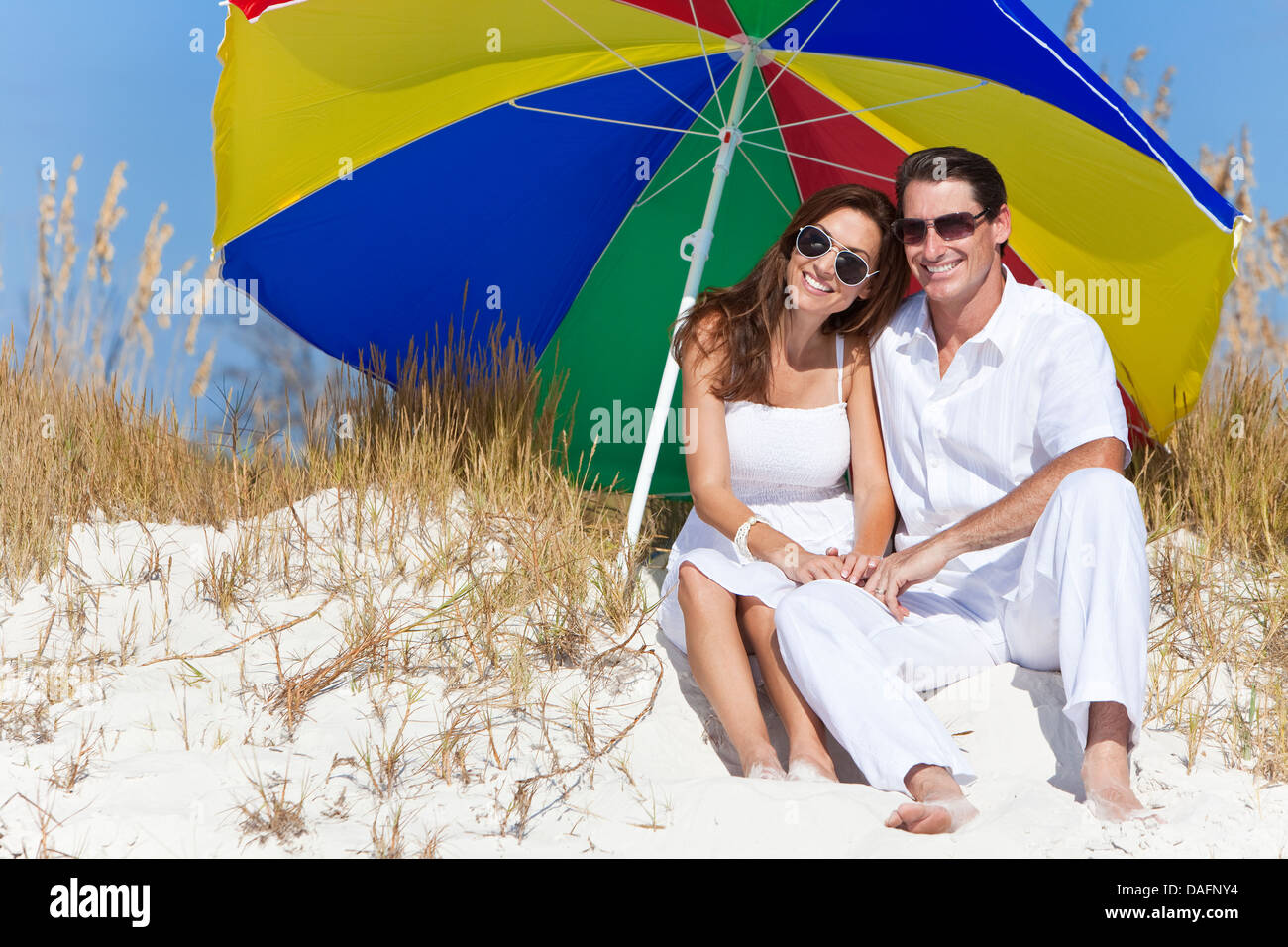 Man and woman romantic couple wearing sunglasses under a multi colored sun umbrella or parasol on a beach Stock Photo