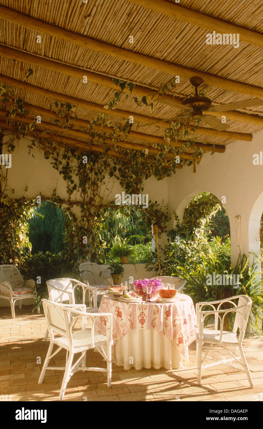 White chairs at table with patterned cloth set for lunch on veranda of ...